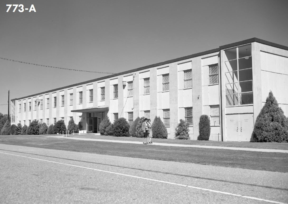 A picture from 1960 of a long building with many windows with two women in business attire standing out front.