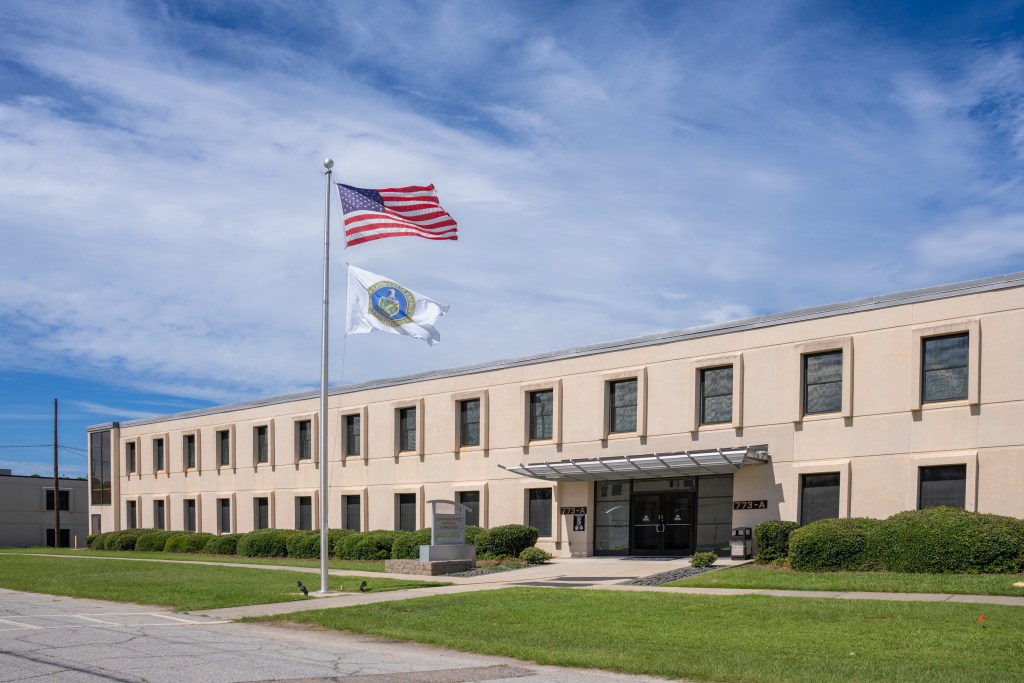 A picture of a long building with many windows, grass, shrubbery, and a flagpole flying the American flag and the Department of Energy flag.