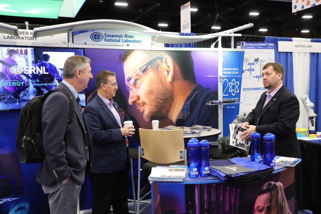 Three men in business suits talking and drinking coffee while standing around a table with SRNL flyers, bags and reusable water bottles.