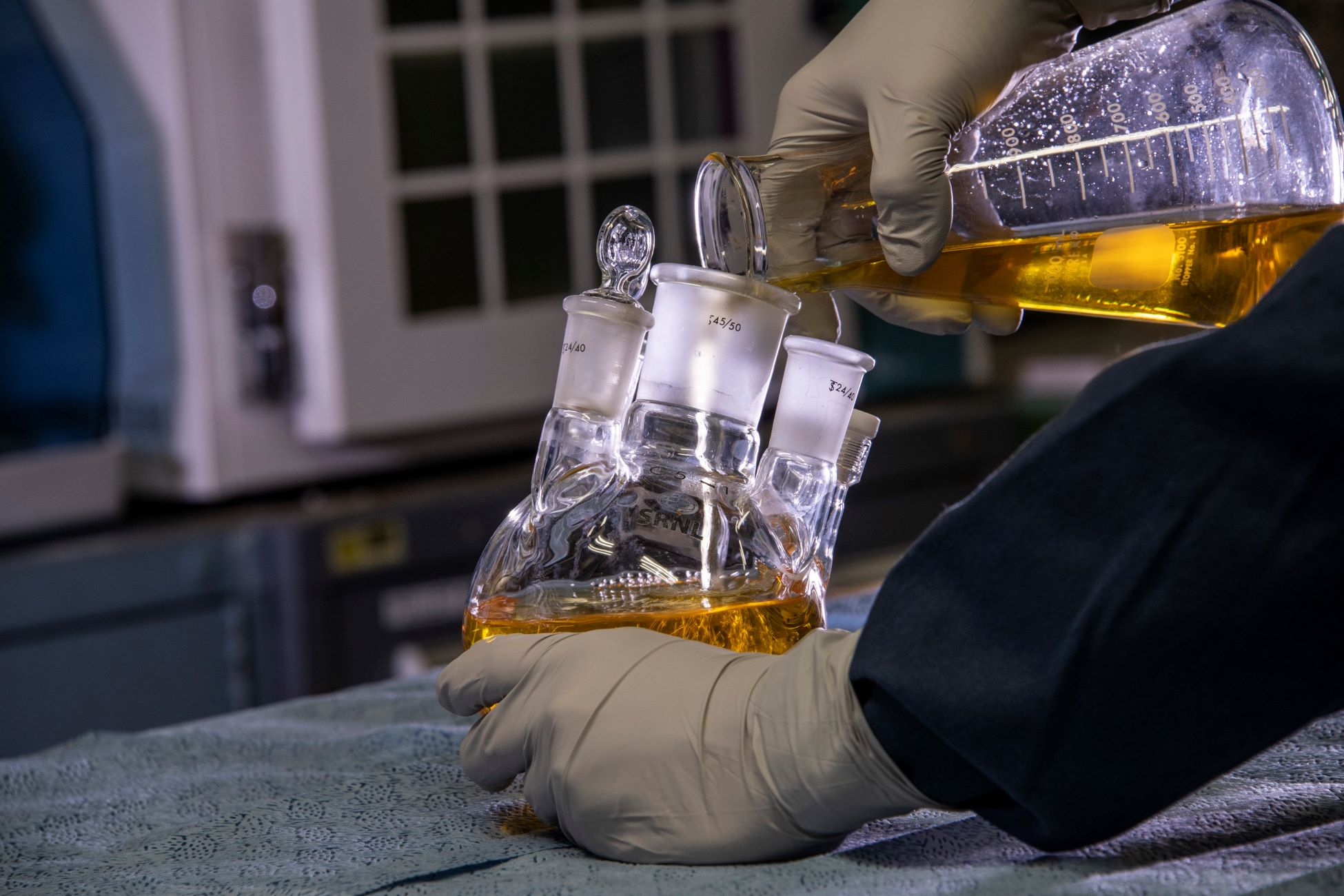 A scientist wearing gloves pouring liquid from flask into a beaker