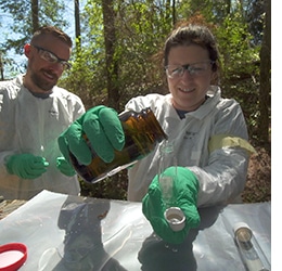 An intern pouring a jar into a vial while another intern watches behind her.